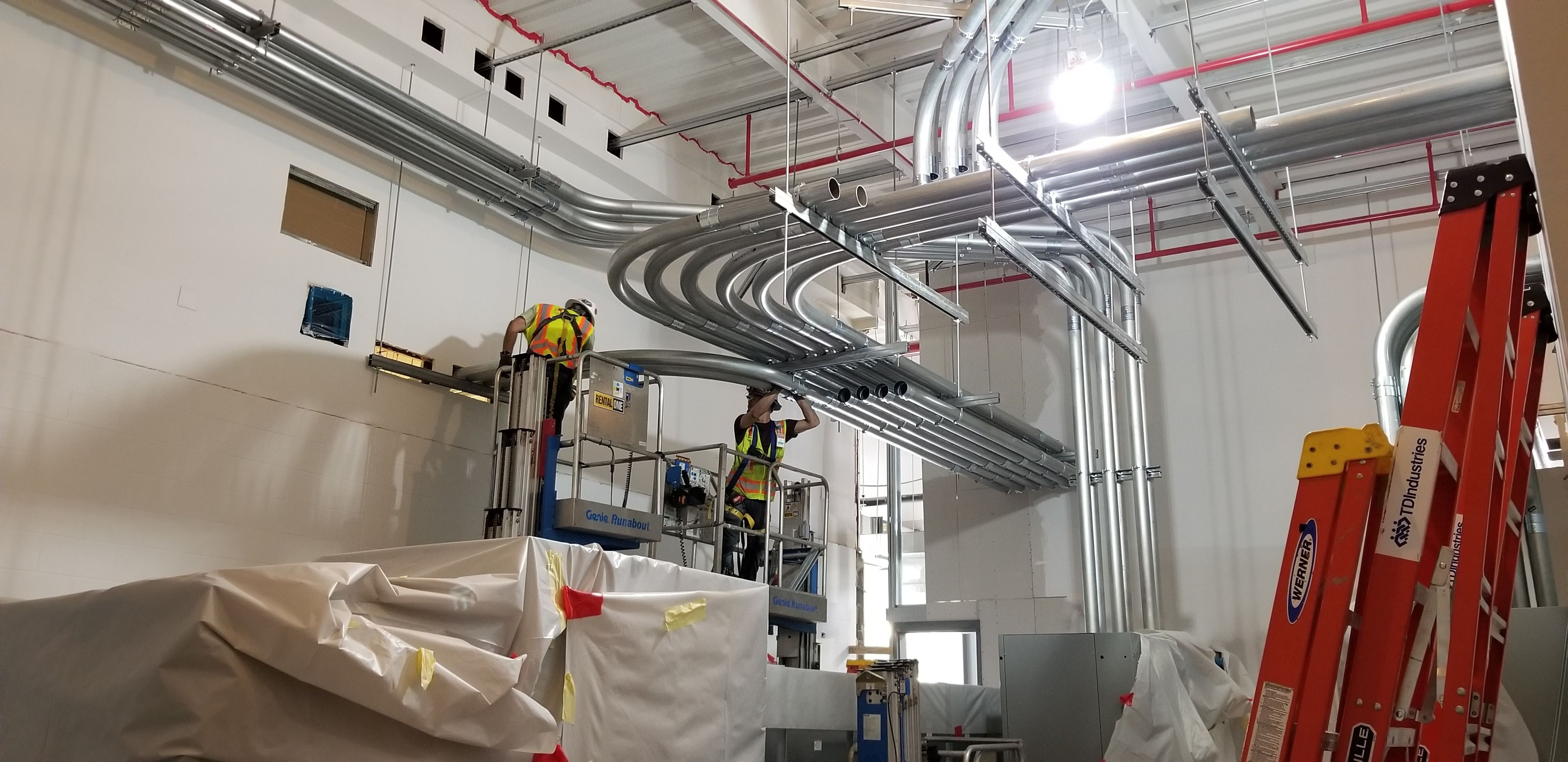 Electricians on lifts installing overhead conduit racks in a commercial facility
